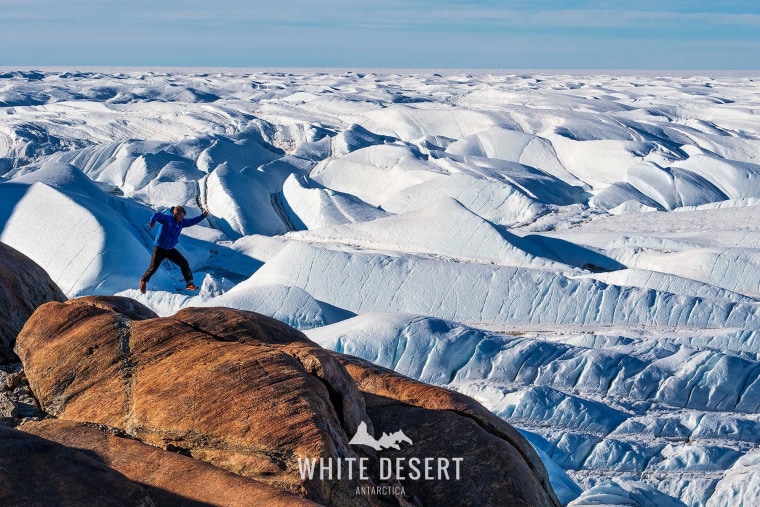 A man jumps on a rock in Antarctica in a blue puffer jacket. Behind him are white glaciers of snow.