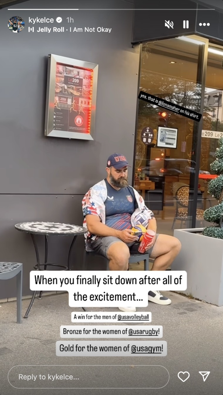 Jason Kelce in American-colored apparel sits outside a cafe and tiredly scrolls on his phone.