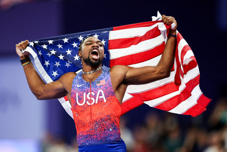 Noah Lyles runs hold and American flag behind him in celebration