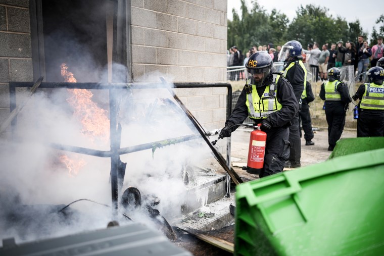 A Riot police officer uses a fire extinguisher on a fire in front of a doorway billowing smoke