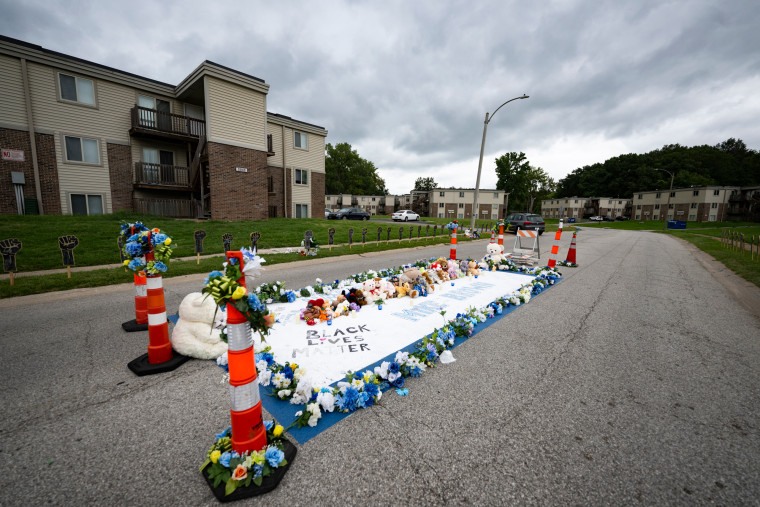 A memorial for Michael Brown in the center of a street near apartment building is laden with flowers, cones, and text that reads "Black Lives Matter" and "Mike Brown"