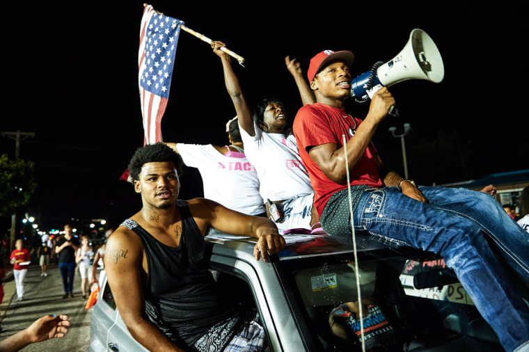 Demonstrators protest on the street, a woman waves an American flag sitting atop a car next to a man holding a megaphone