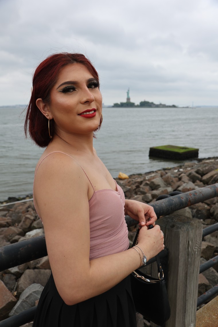 Michelle Carranza smiles outside, the statue of Liberty can be seen across the water in the distance