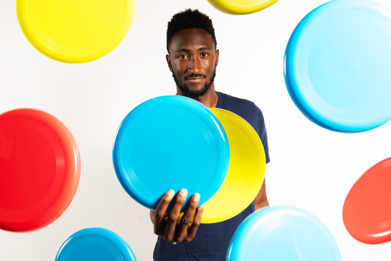 Marques Brownlee holding two frisbees with multicolored frisbees in the air around him.