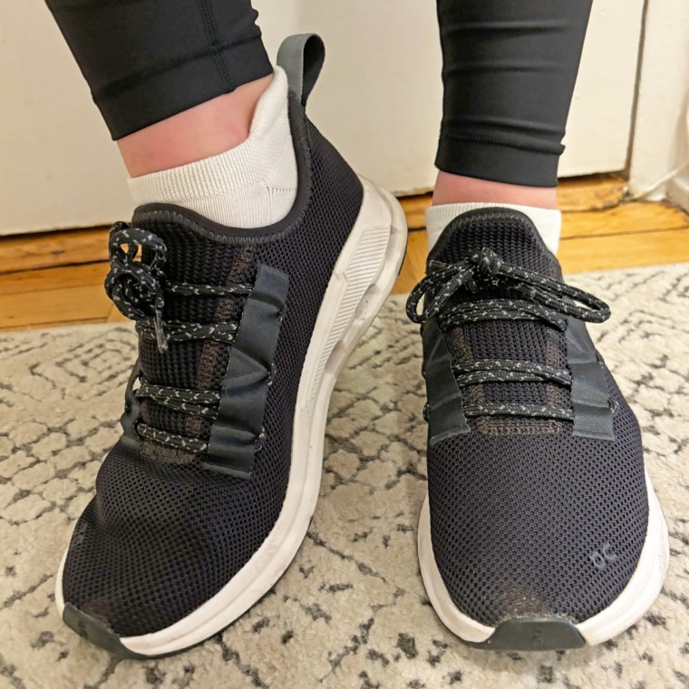 A woman wearing a black pair of On Cloudeasy walking shoes while she stands inside on a carpet.