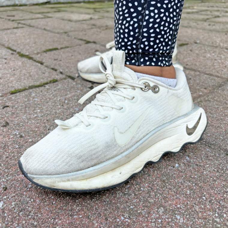 A white Nike Motiva walking sneaker on a women's foot as she stands outside on a stone floor.