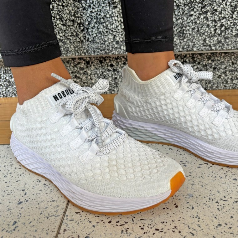 A woman wearing white Nobull Aspire sneakers while standing inside on a tile and wood floor.