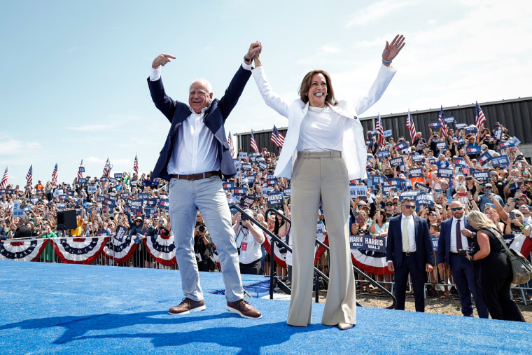 TIm Walz and Kamala Harris on stage outside, hold hands and raise their arms in celebration