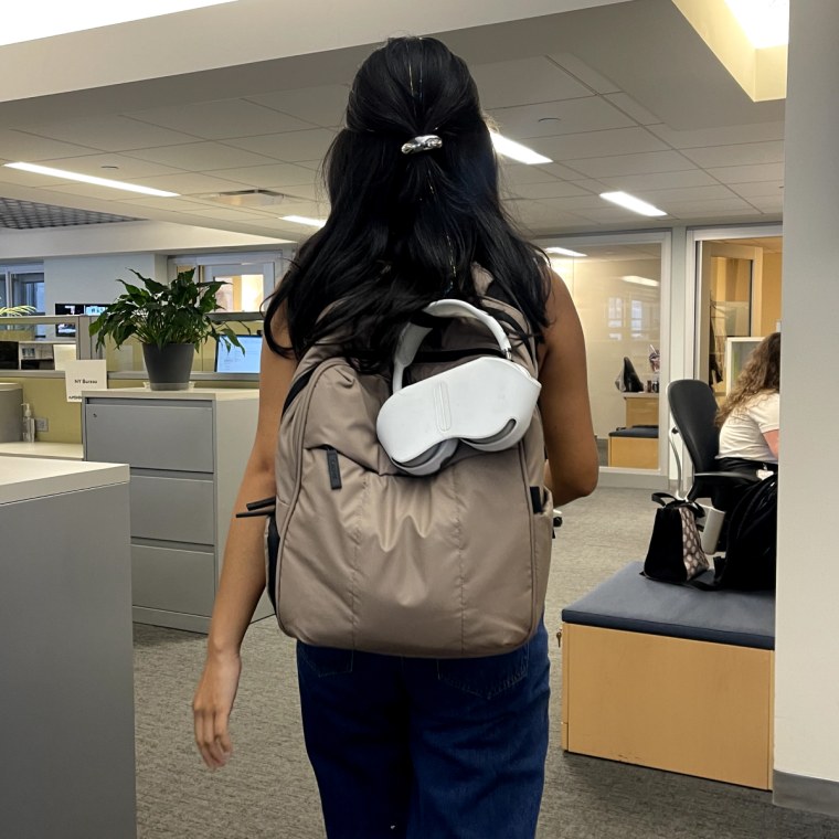 The woman wears a brown backpack in the office with a white AirPod Max attached to the hanging loop of the backpack