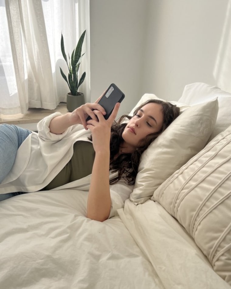 Woman lays on her bed with her head on a Blissy Silk Pillowcase as she scrolls on her phone