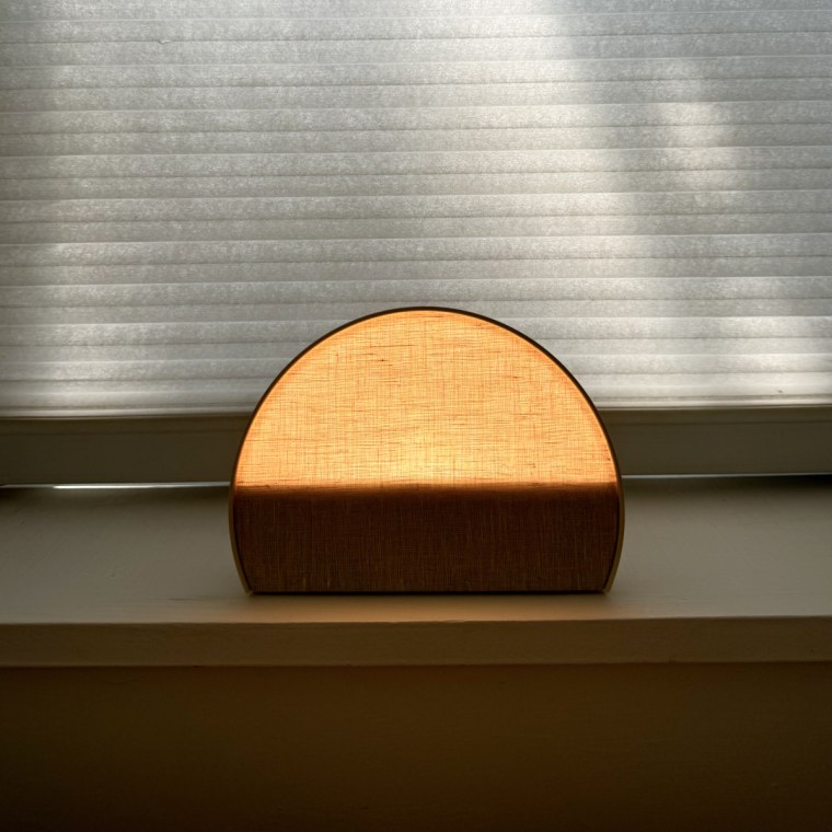 A Hatch Restore 2 alarm clock sits on top of a white windowsill, shining a bright peach color.