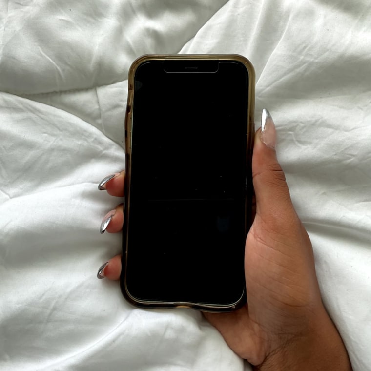 A right hand with silver tipped nails holding an iPhone 12 Plus against a white duvet cover background