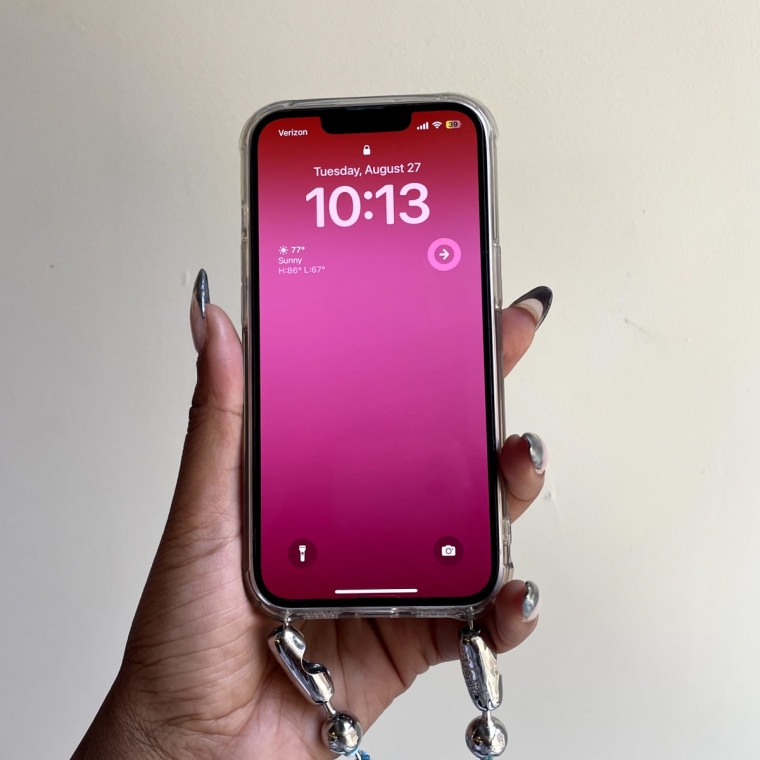 A left hand with silver tipped nails holding an iPhone with a pink gradient lockscreen and the time 10:13 AM against a beige wall.