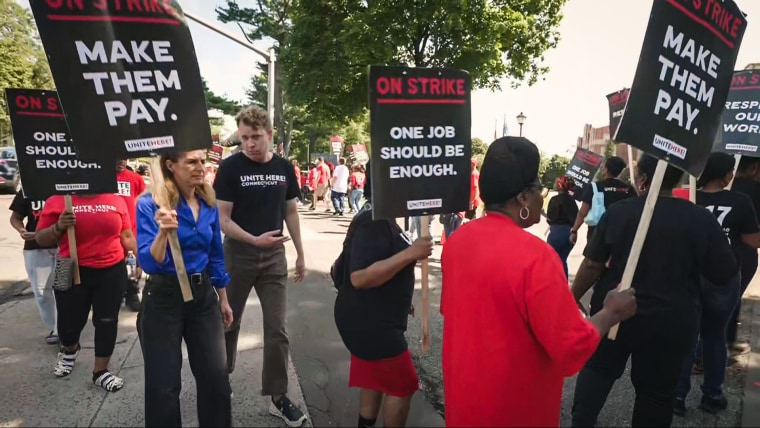 People hold signs saying "Make Them Pay." and "One Job Should Be Enough."