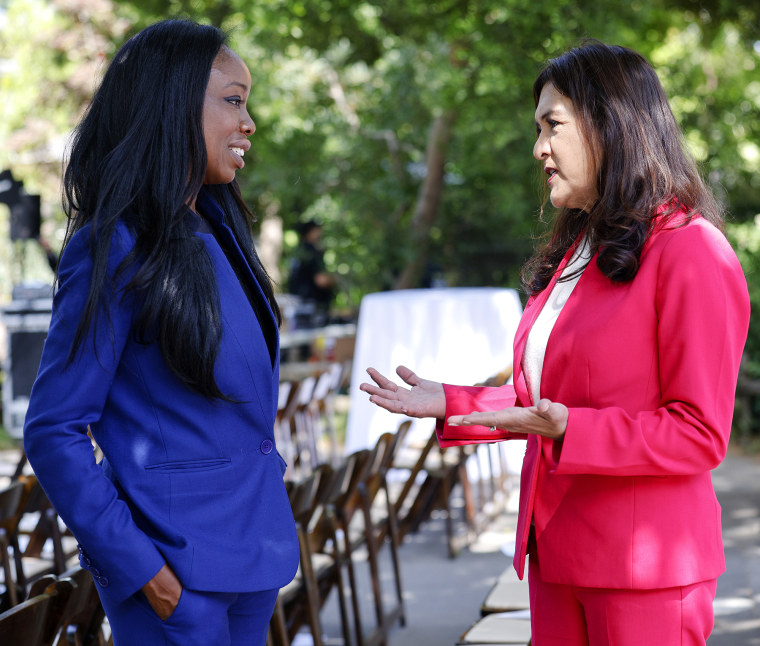 Woman in blue pantsuit and woman in pink pantsuit talk with each other outdoors