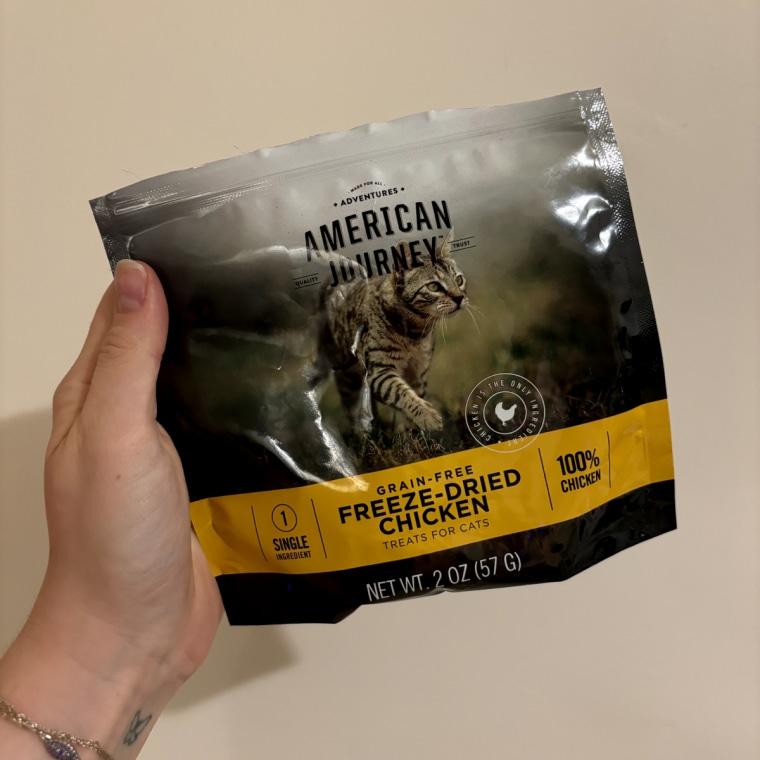 A woman’s hand holding a bag of American Journey Grain-Free Freeze-Dried Chicken Treats against a white wall.