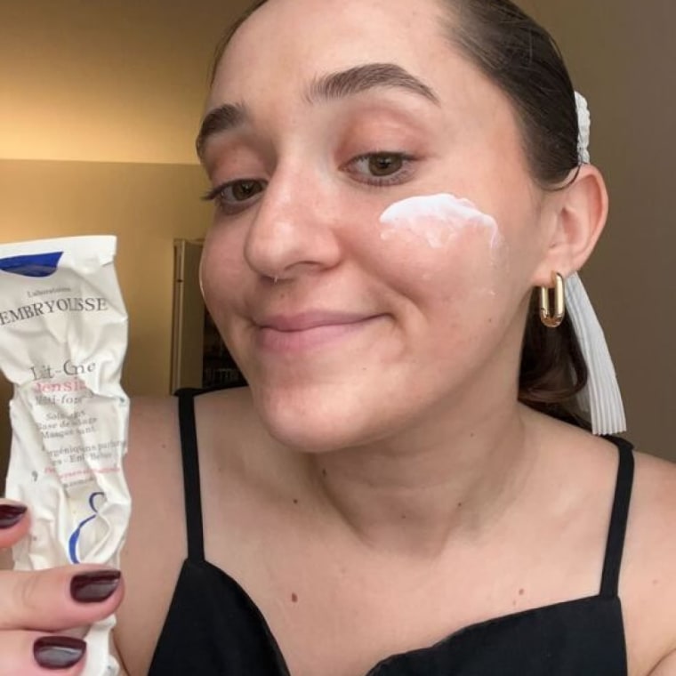 Woman holds an almost empty tube of Embroylisse Lait Crème Concentré and has a swatch of the moisturizer on her right cheek.