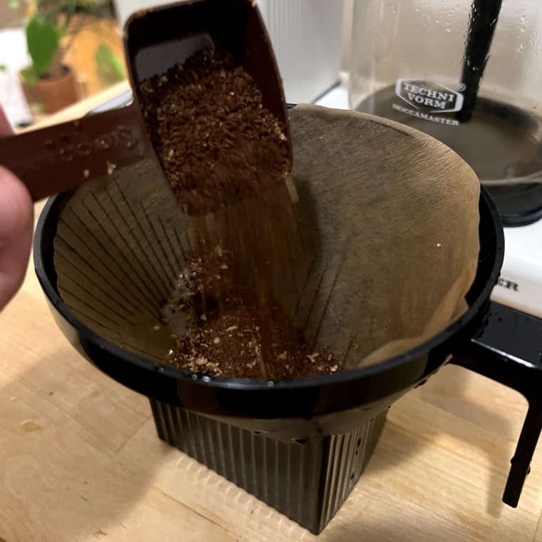 Hand scooping coffee beans into a paper filter inside a black brew basket