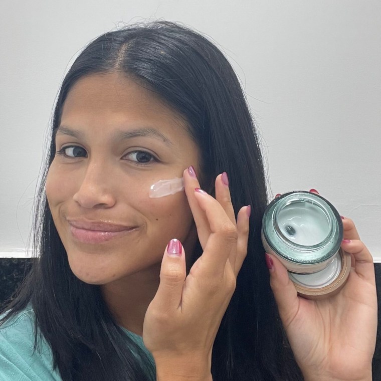 Woman holds an open jar of the moisturizer in her hand and swatches it on to her cheek using the other hand.