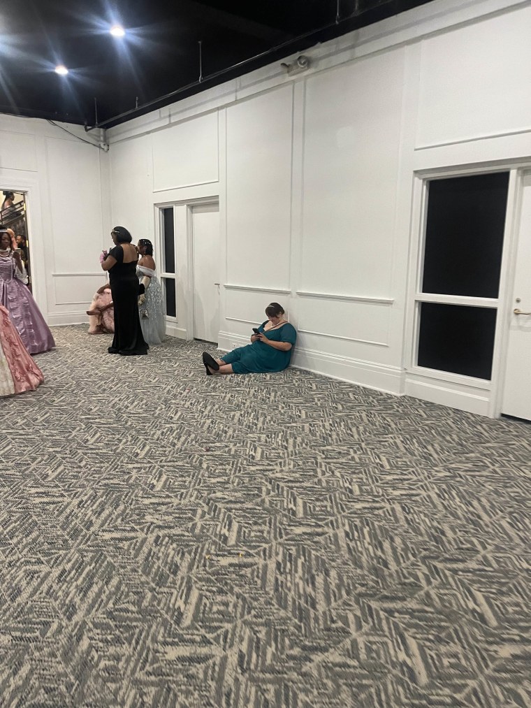 A woman in a teal dress sits on the floor of a ballroom on her phone.