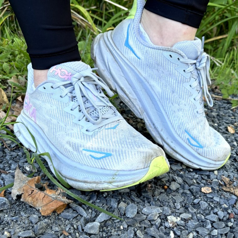 Woman’s feet standing on a gravel road in front of grass, modeling a periwinkle pair of Hoka Clifton 9 sneakers.