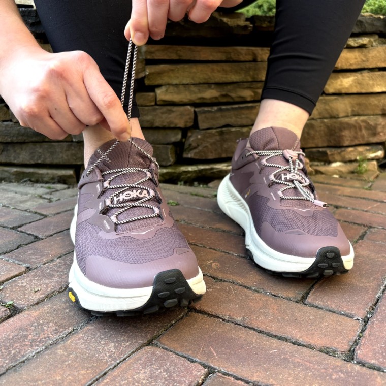 A woman sitting against a stone wall, pulling the elastic laces tighter on the Hoka Transport  sneaker.