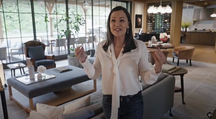 Jennifer Garner in jeans and a white shirt talks to the camera in front of her living room.