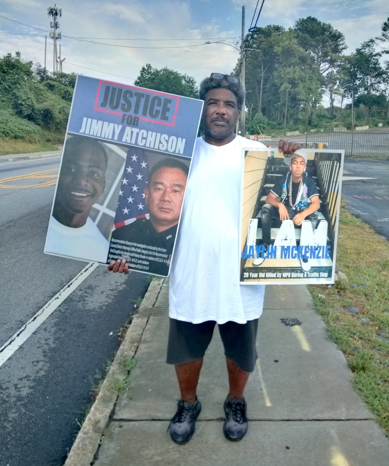 Black man standing on sidewalk holding poster that reads "Justice for Jimmy Atchison" and another that reads "Jaylin McKenzie"