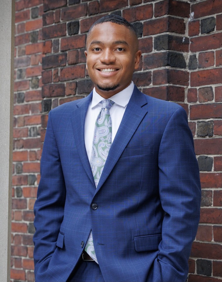 Young Black man in suit smiling, standing against brick wall 