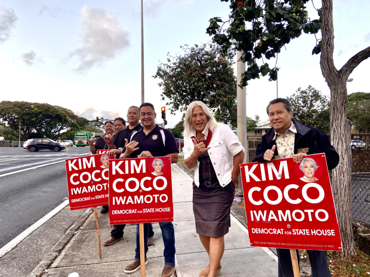 Foto del sujeto Kim Coco Iwamoto de pie en una acera con otras 5 personas que cada una de las antiguas campañas rojas firma con su nombre. 