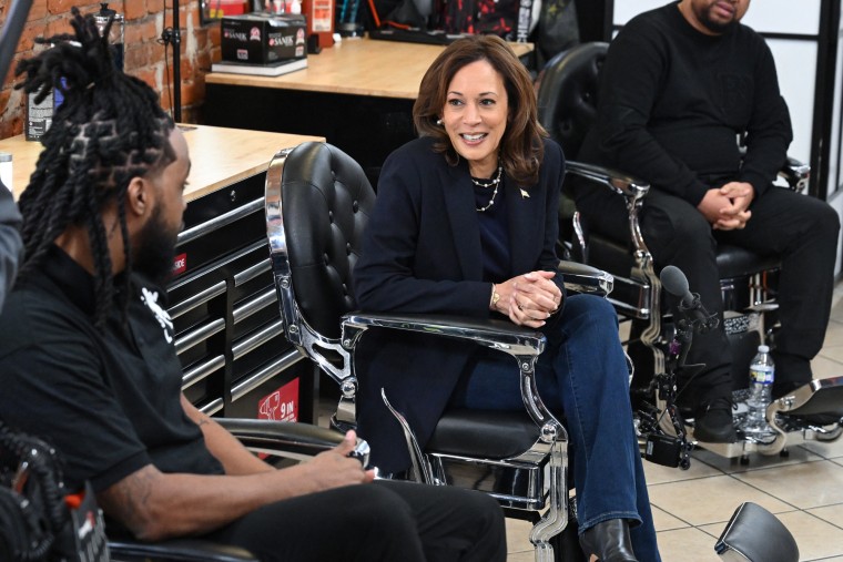 Kamala Harris sits in a chair in a barber shop and speaks to someone sitting next to her