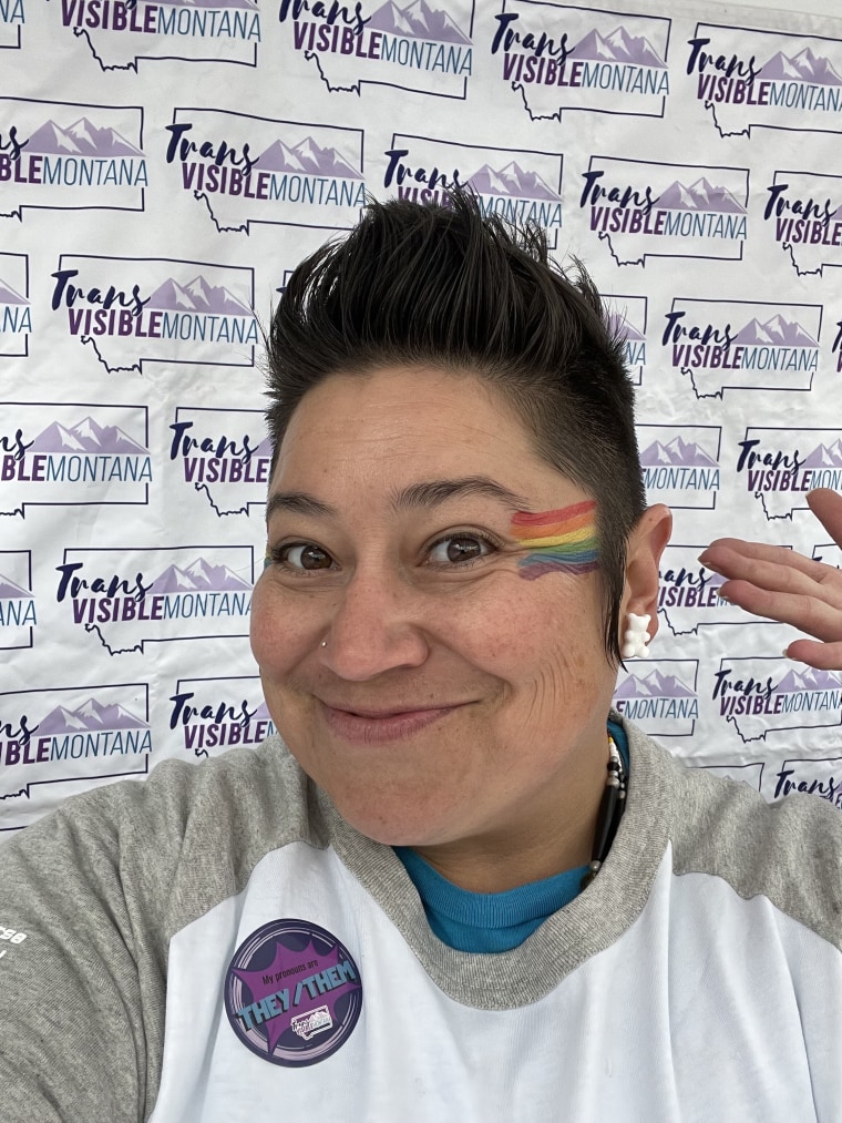 Close-up of a person smiling with a pride flag painted on their face, against a backdrop that reads "Trans Visible Montana" 