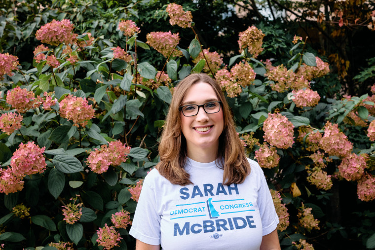 Sarah McBride stands in front of a bush with pink flowers and smiles