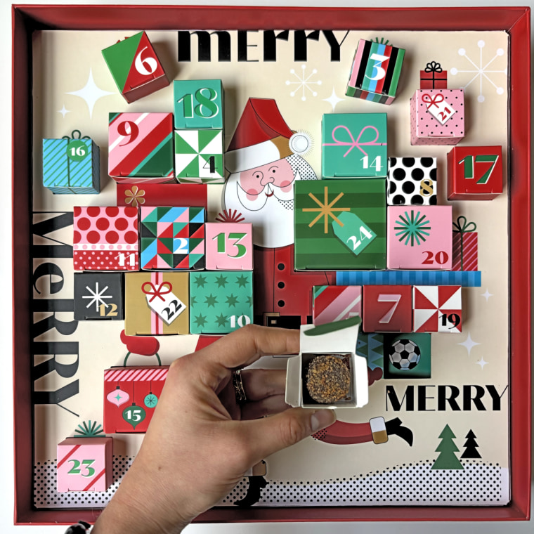 A woman’s hand holding a box with a chocolate truffle inside of it in front of the Christopher Elbow Advent calendar.