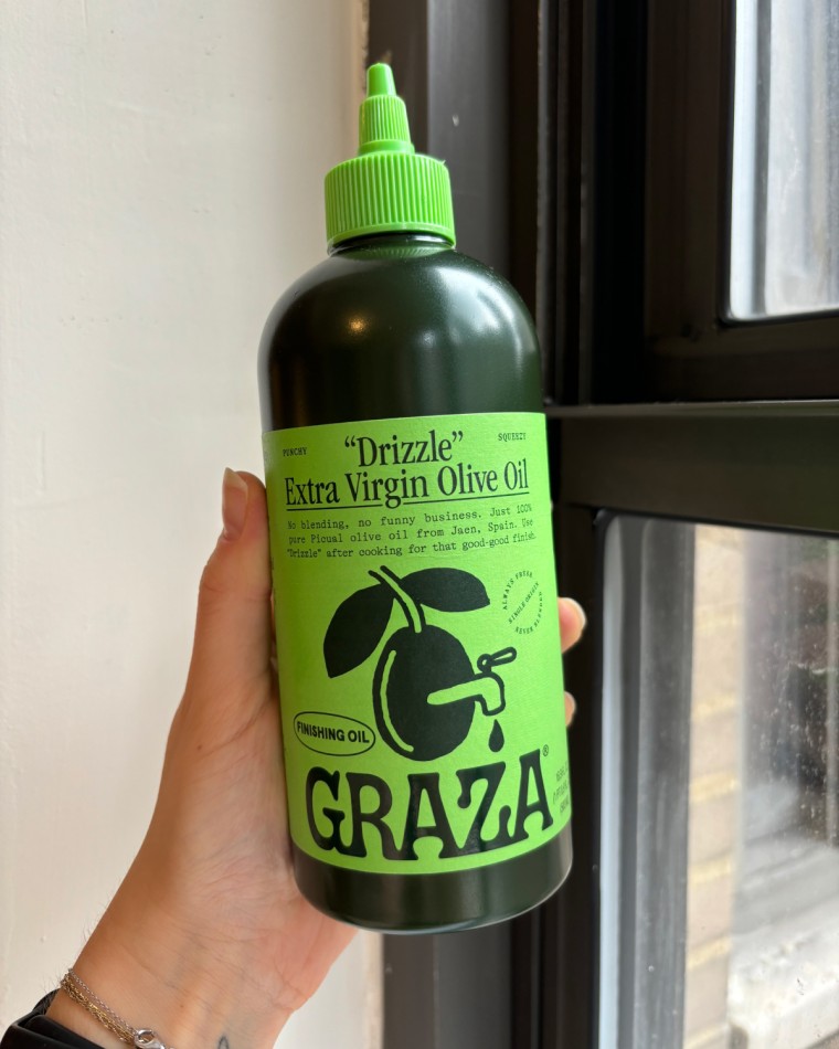 Woman’s hand holding a squeeze bottle of Graza’s Drizzle Extra Virgin Olive Oil in front of a window.