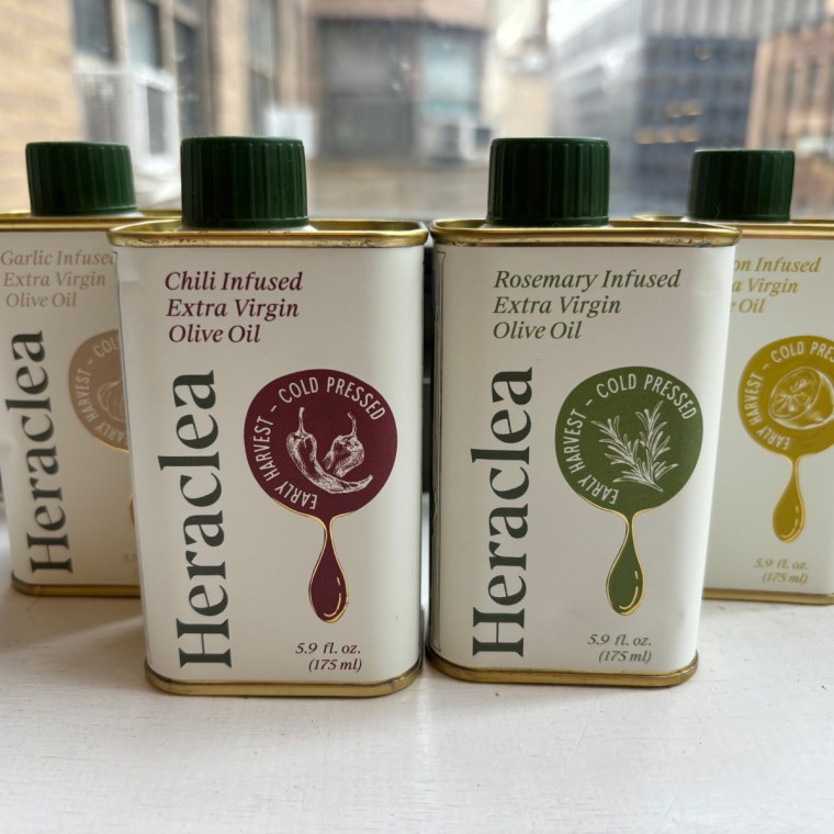 Four bottles of Heraclea’s flavored olive oils lined up in front of a window. From left to right: garlic, chili, rosemary and lemon.