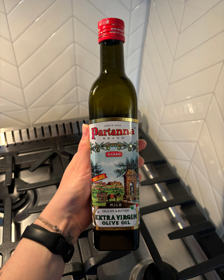 A woman’s hand holding a bottle of Partanna olive oil in front of a gas stovetop and white backsplash.