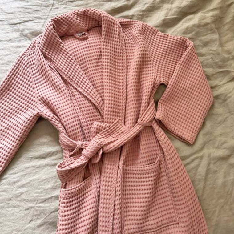 A rose-colored waffle robe lays flat and ties on a beige bed.