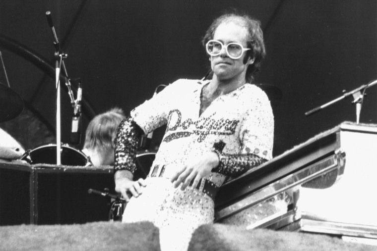 Elton John leans against a piano on stage