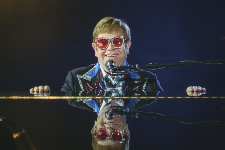 Elton John smiles while sitting behind a piano