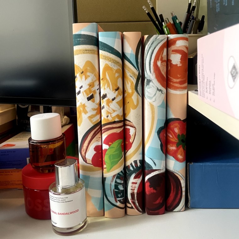 Five books covered in food-themed Juniper book covers, with the bottom right corner of a computer screen visible to the left of the covers. The covers are beside two bottles of fragrance and a jar of body cream.
