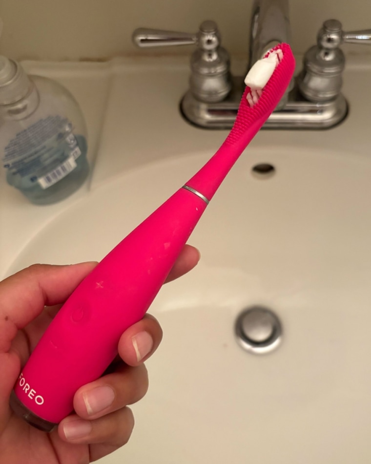 A woman’s hand holding a hot pink Foreo Issa 3 Rechargeable Electric Toothbrush with toothpaste on it over a white bathroom sink.