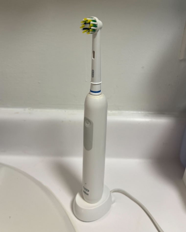 A Oral-B Pro 1000 Electric Toothbrush standing up in its holder and sitting on a white bathroom sink.