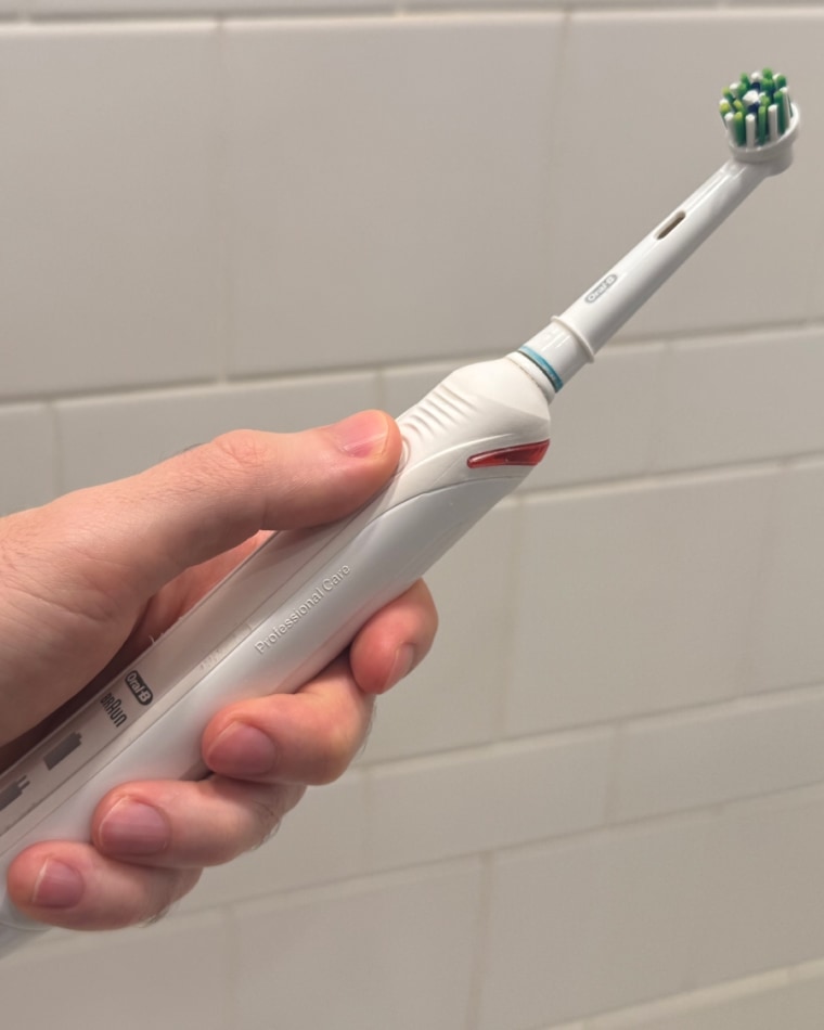 A man’s hand holding the Oral-B Smart 1500 Electric Toothbrush against a white tile wall.