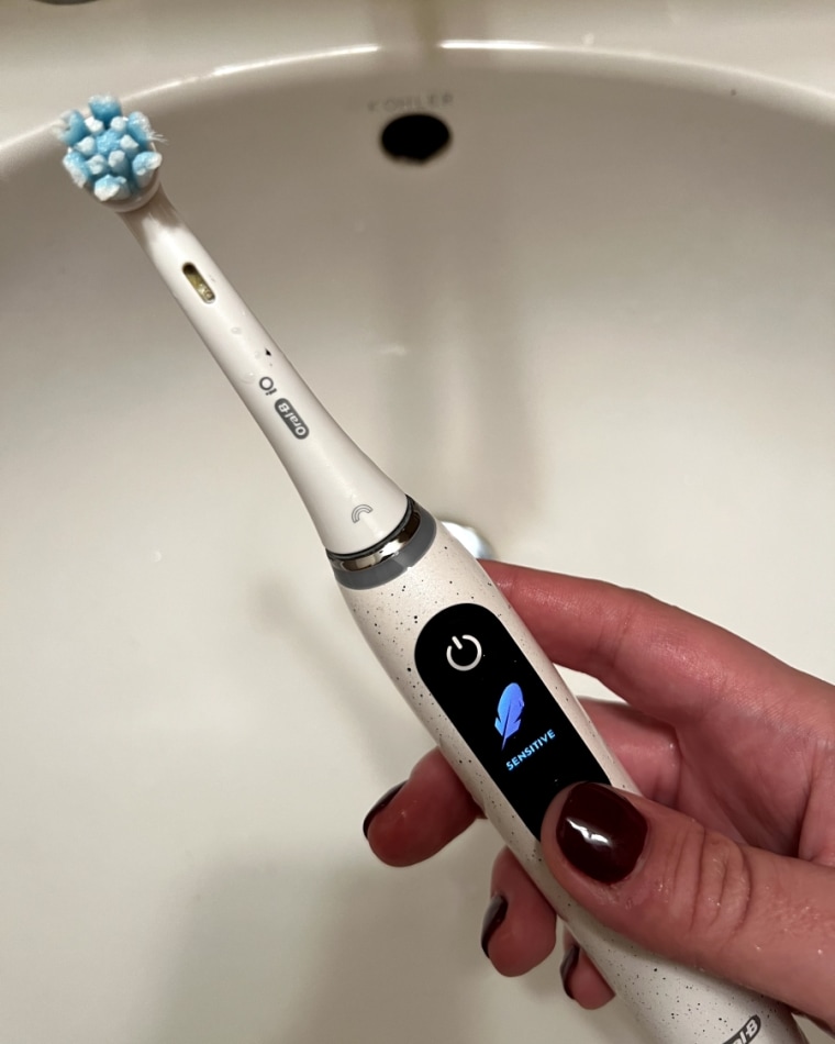 A woman’s hand holding the Oral-B iO Series 10 Electric Toothbrush over a bathroom sink with the word “sensitive” displayed on the handle’s screen.