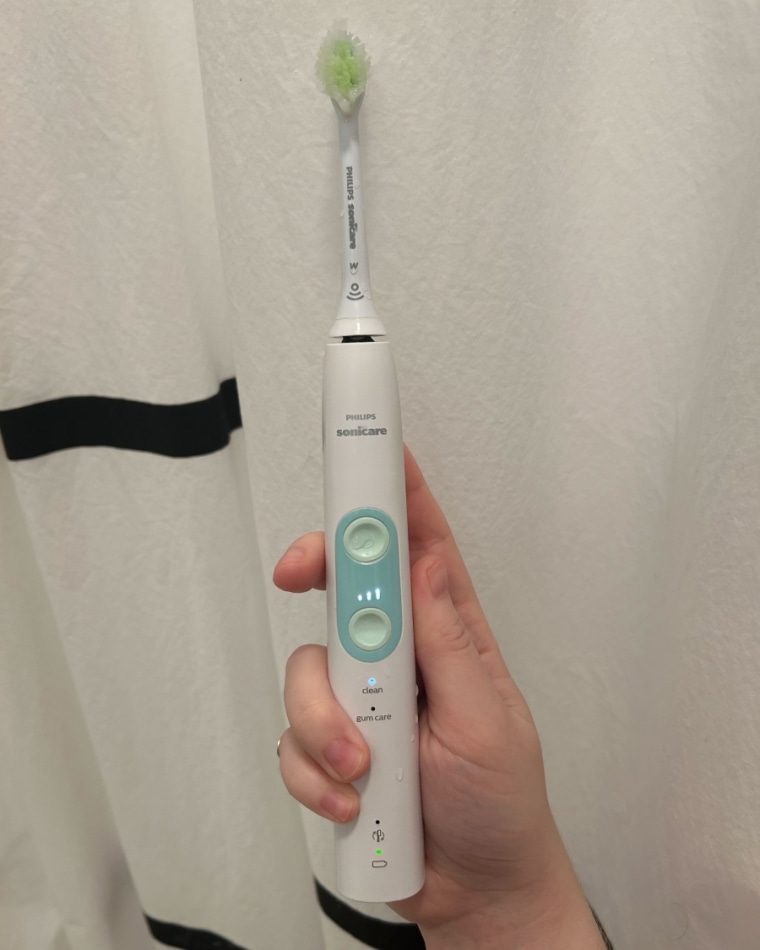 A woman’s hand holding the Philips Sonicare ProtectiveClean 5100 Electric Toothbrush against a black and white shower curtain.