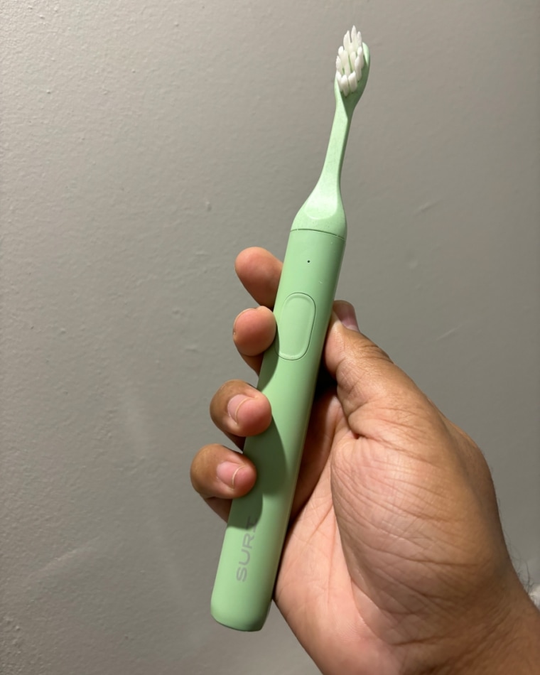 A man’s hand holding the green Suri Sonic Toothbrush against a gray wall.