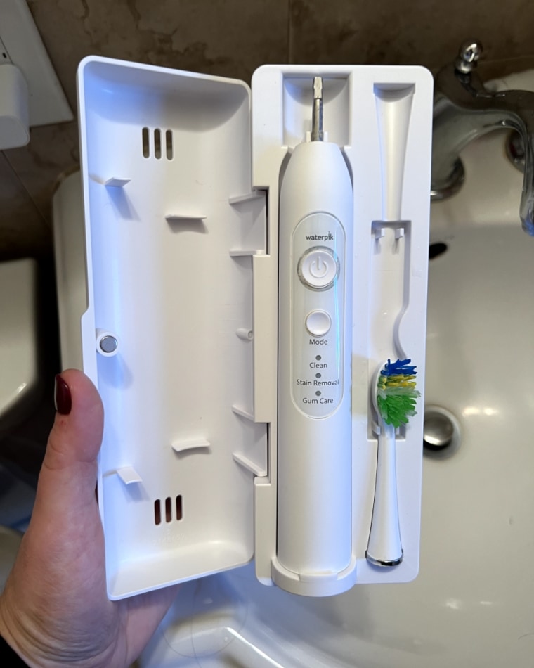 A woman’s hand holding the Waterpik Sensonic Electric Toothbrush in its travel case over a sink in a bathroom.