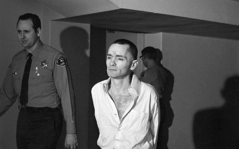 Charles Manson, with a short haircut and a swastika mark on his forehead, is escorted through an LA courthouse by sheriff's deputies.