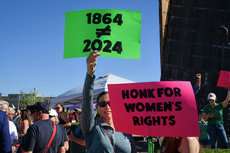 A woman holds a green sign that says "1864 does not equal 2024", and a pink sign that says "Honk for women's rights"